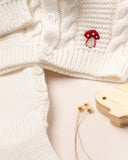 Na imagem, vemos detalhes do sofisticado bordado exclusivo de cogumelo vermelho no casaquinho na cor off white. 
